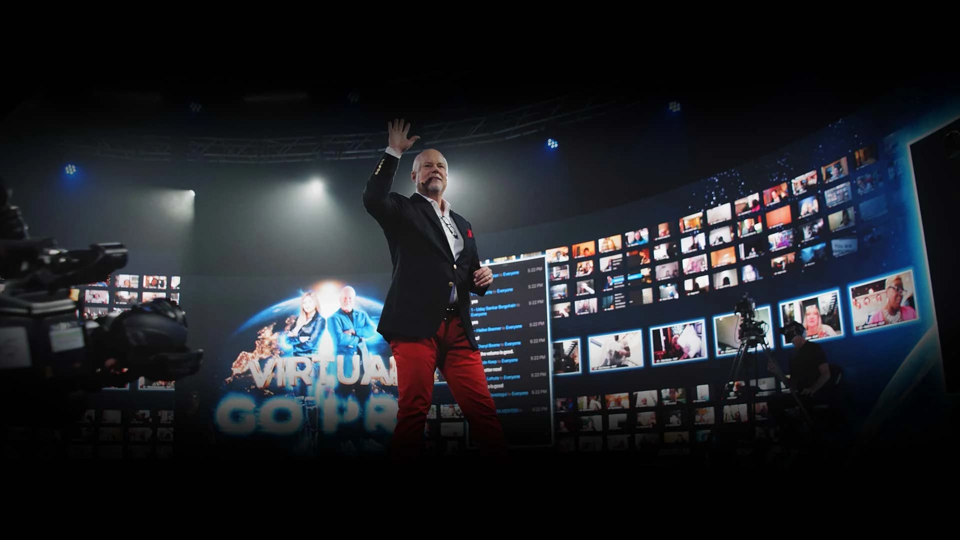 A presenter on the 360° stage waving to the audience, hundreds of remote attendees live on the wraparound LED wall behind him.