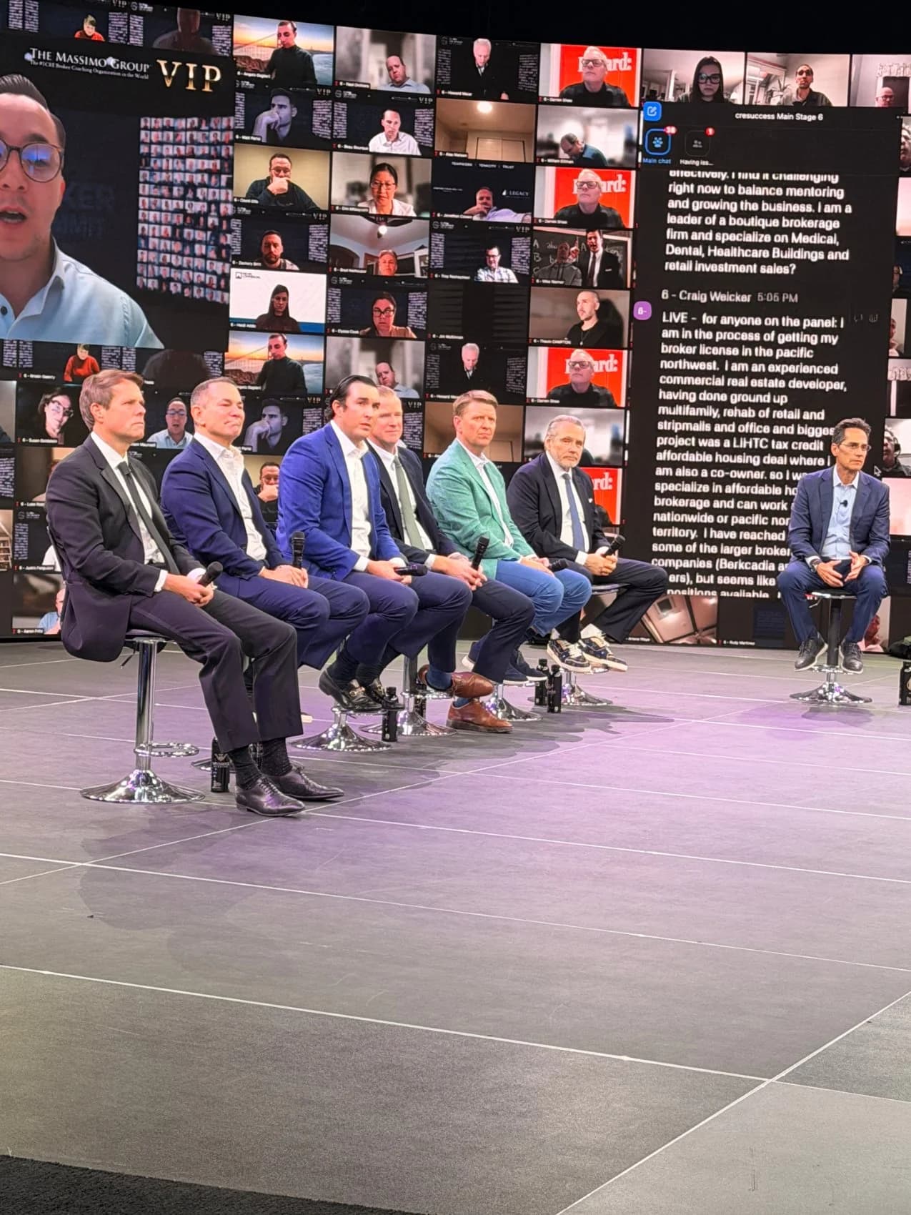 A live broker panel of seven seated speakers on the studio stage, the audience wall of remote attendees rising behind them.