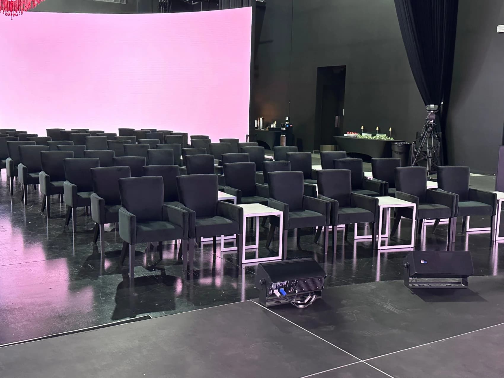 Rows of black lounge armchairs with side tables facing the curved LED wall washed in pink, set for an intimate session.