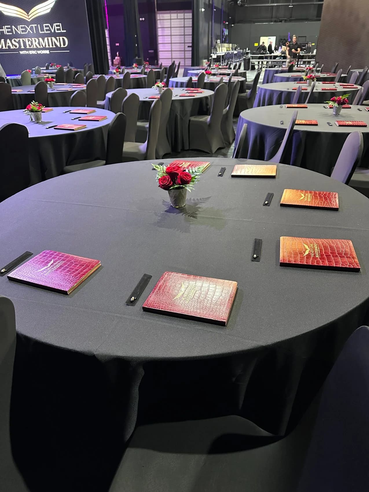 Round banquet tables dressed in charcoal with red roses and gold-edged leather journals for The Next Level Mastermind.