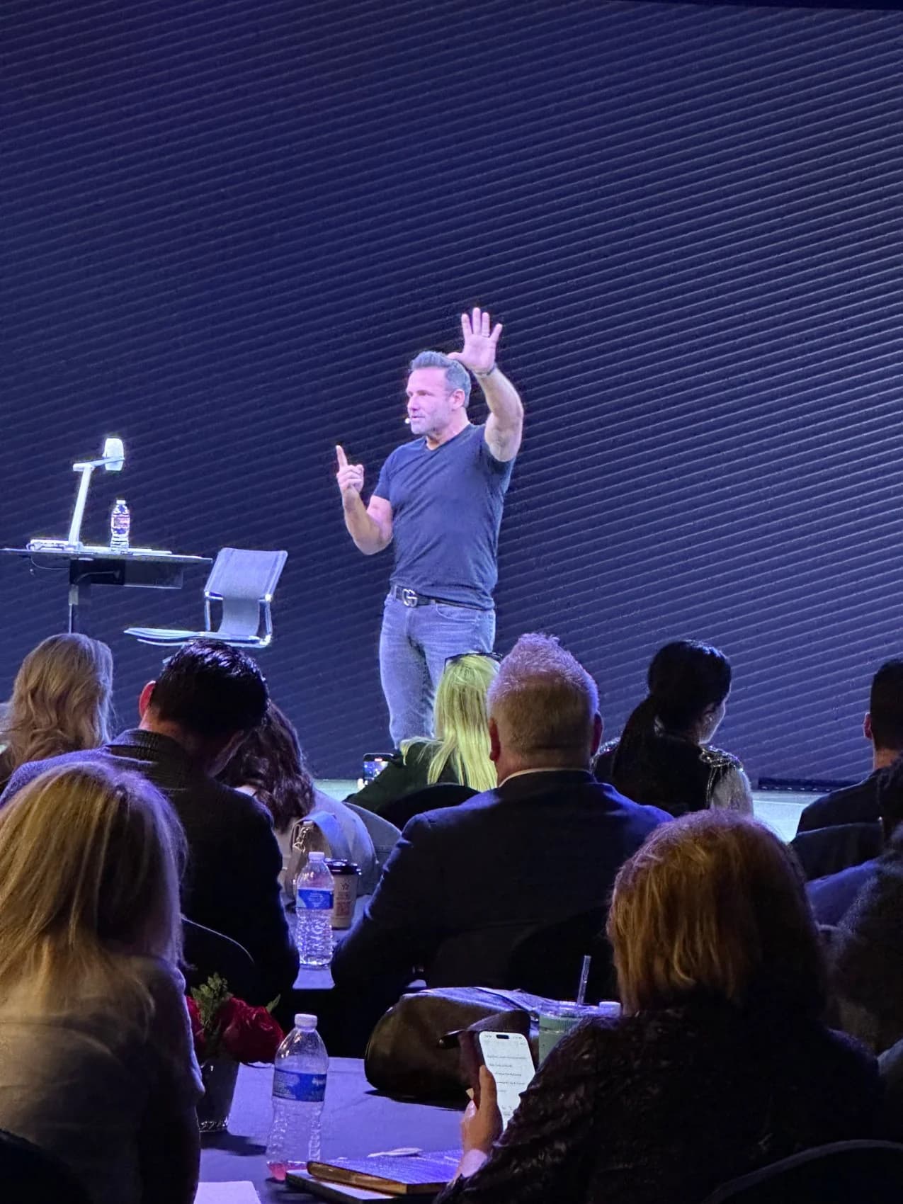 A speaker on stage in a t-shirt addressing a seated audience at The Next Level Mastermind, arm raised mid-point.