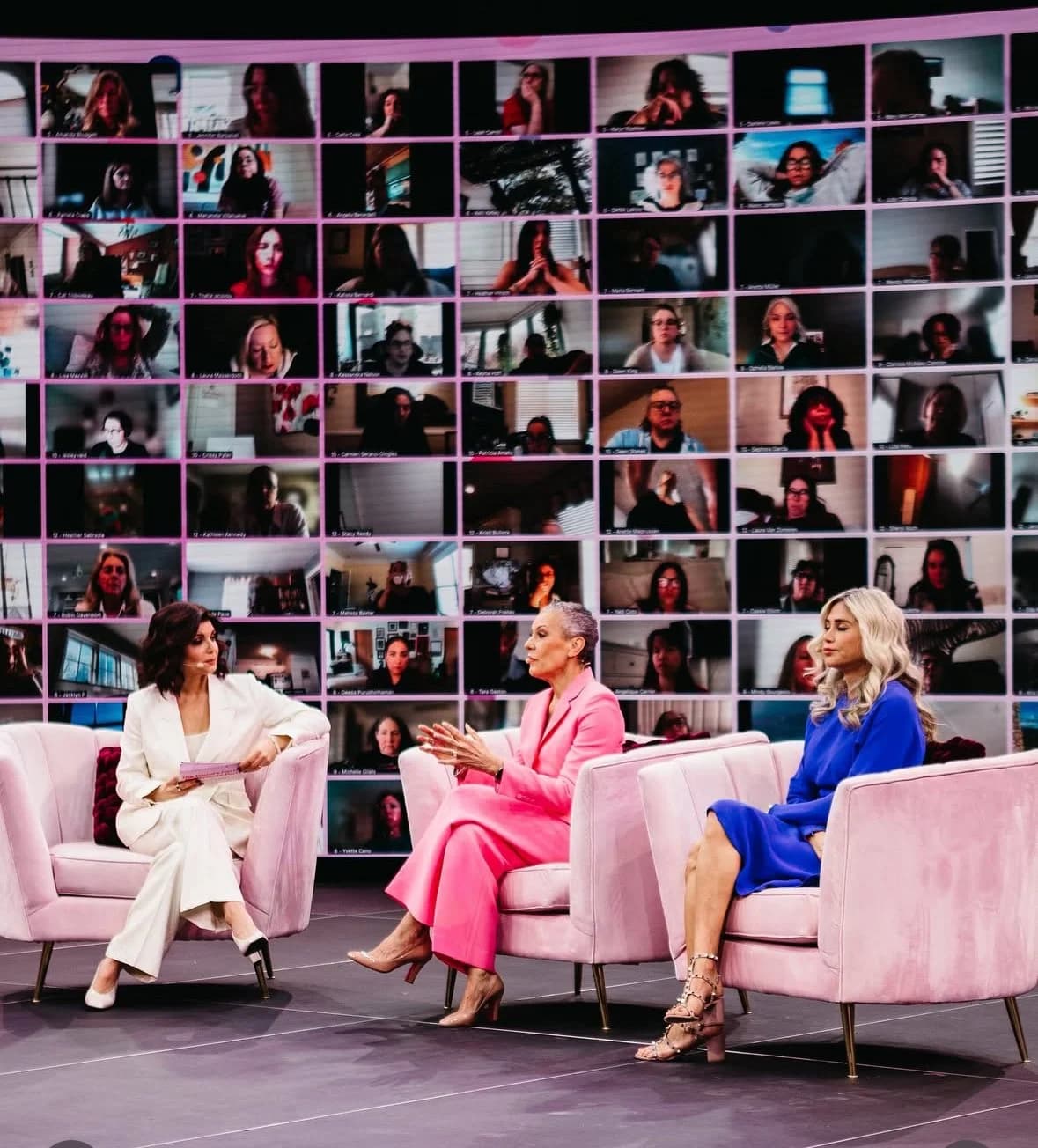 Three women in a moderated panel on pink velvet chairs, a grid of remote attendees filling the LED wall behind them.