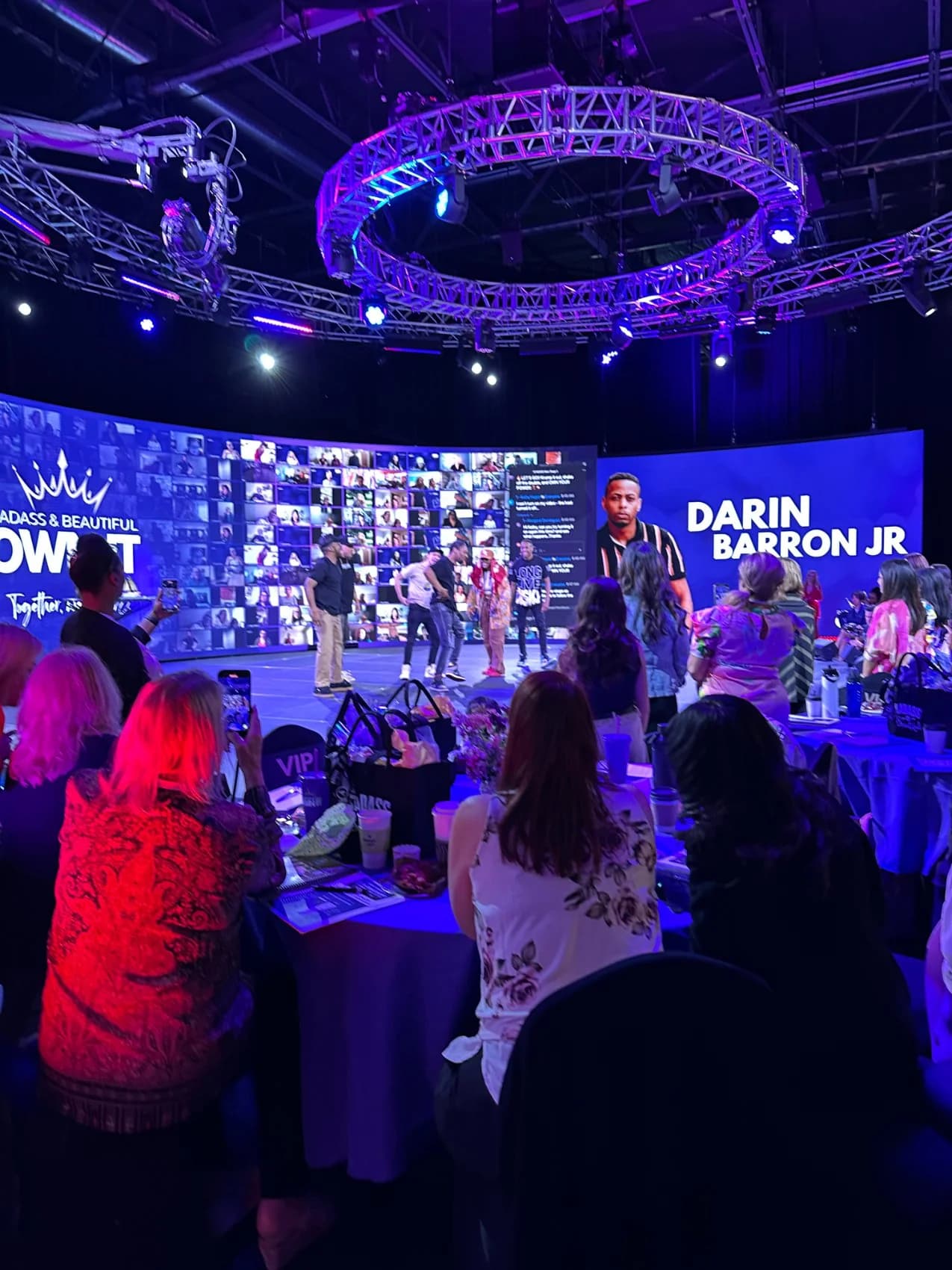 A packed hybrid audience under a circular truss watching hosts on a blue-lit stage, the LED wall showing remote attendees and speaker Darin Barron Jr.