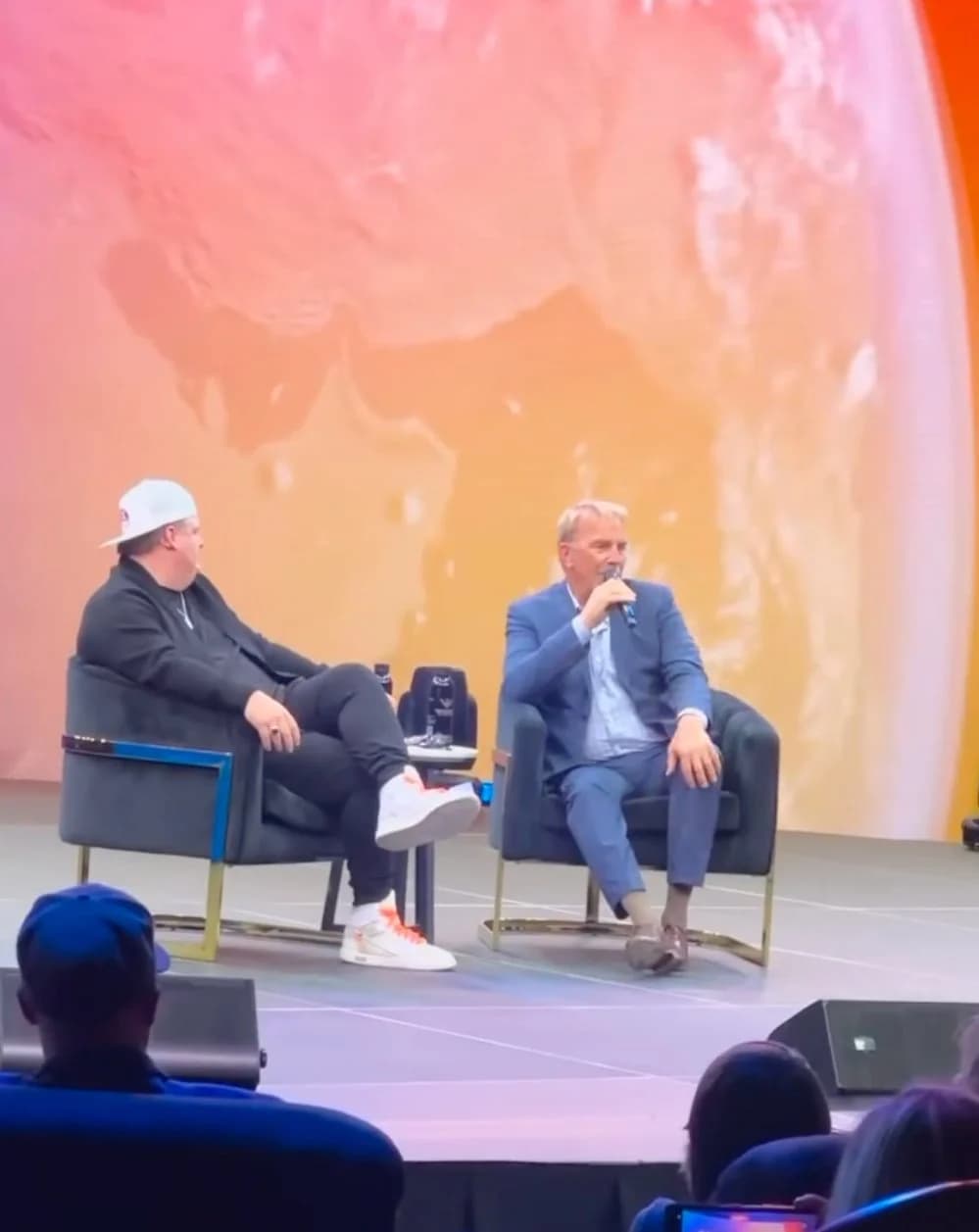 A fireside chat between a host and guest seated on stage in front of a warm orange LED backdrop.