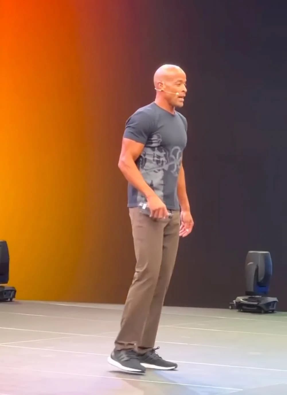 A keynote speaker mid-talk on stage against an orange-to-black lit backdrop.
