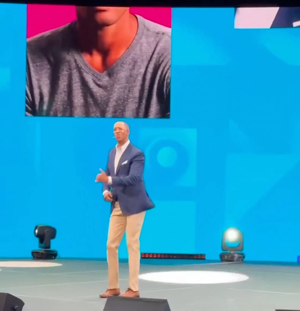 A keynote speaker on a blue-lit stage with a remote attendee pulled up large on the LED wall above him.