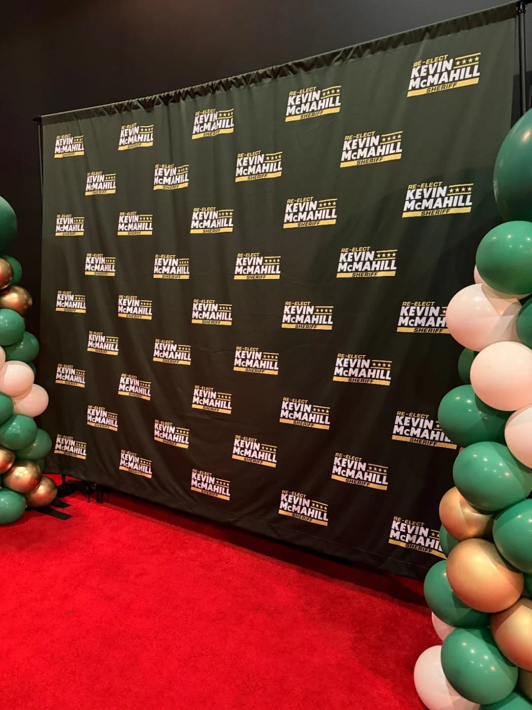 A step-and-repeat campaign backdrop and red carpet framed by green and gold balloon arches at the event entrance.