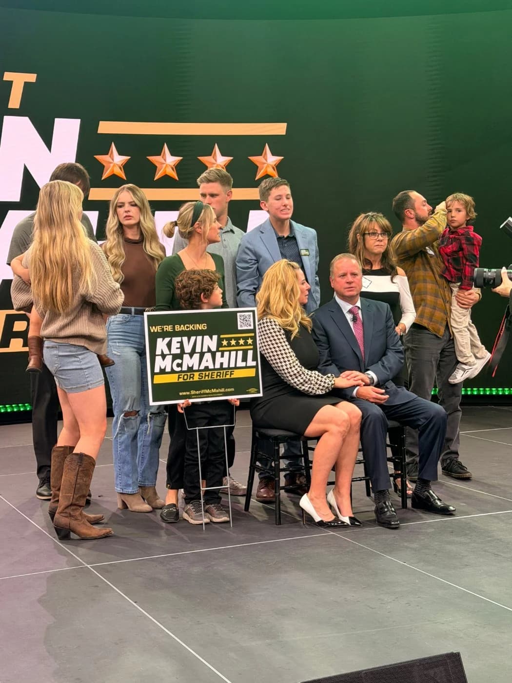 The candidate and his family gathered on stage for photos with a campaign yard sign, the branded LED backdrop behind them.