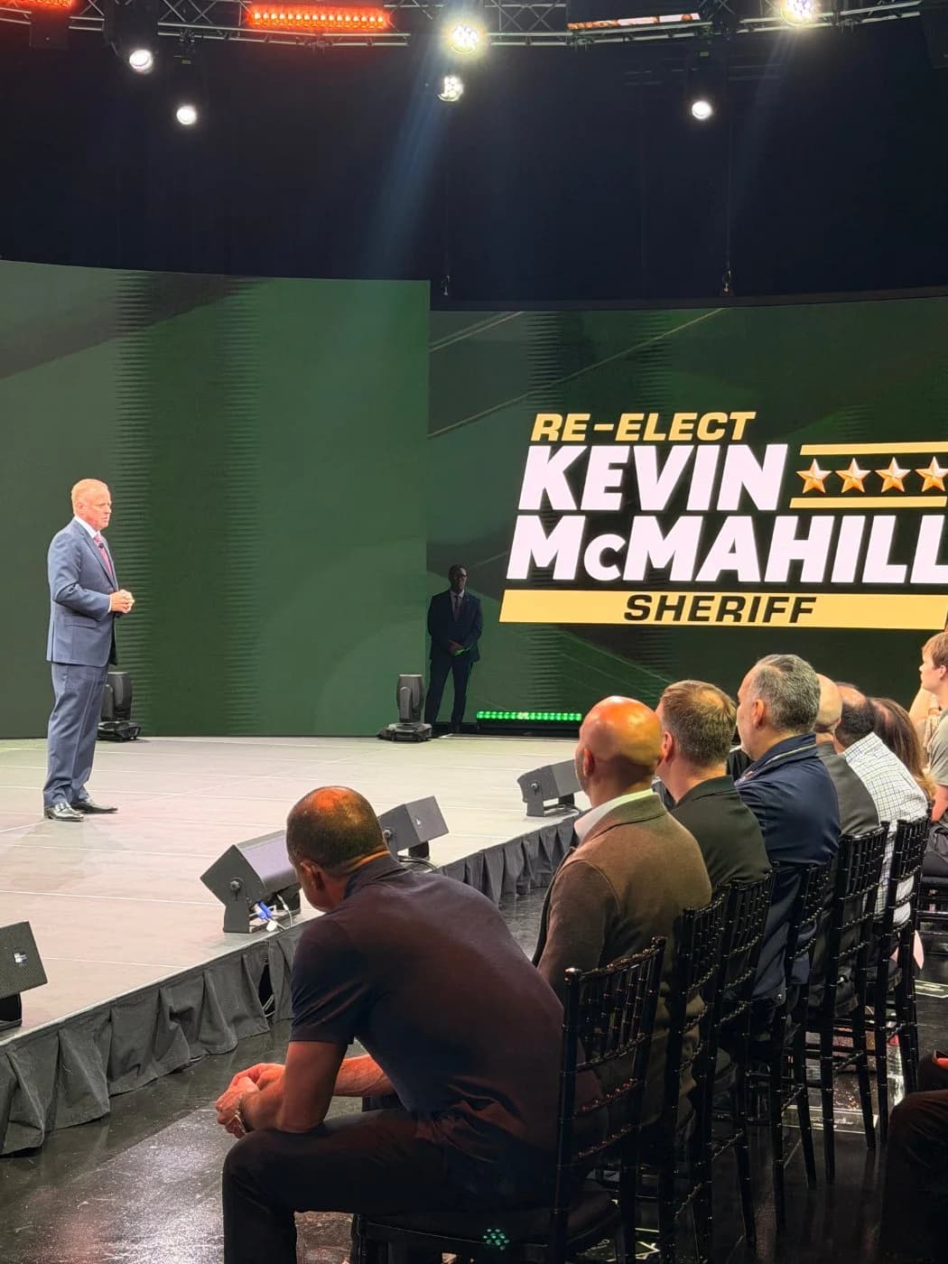 A candidate addressing a seated audience on the 360° stage, a green Re-Elect Kevin McMahill Sheriff graphic filling the LED wall behind him.