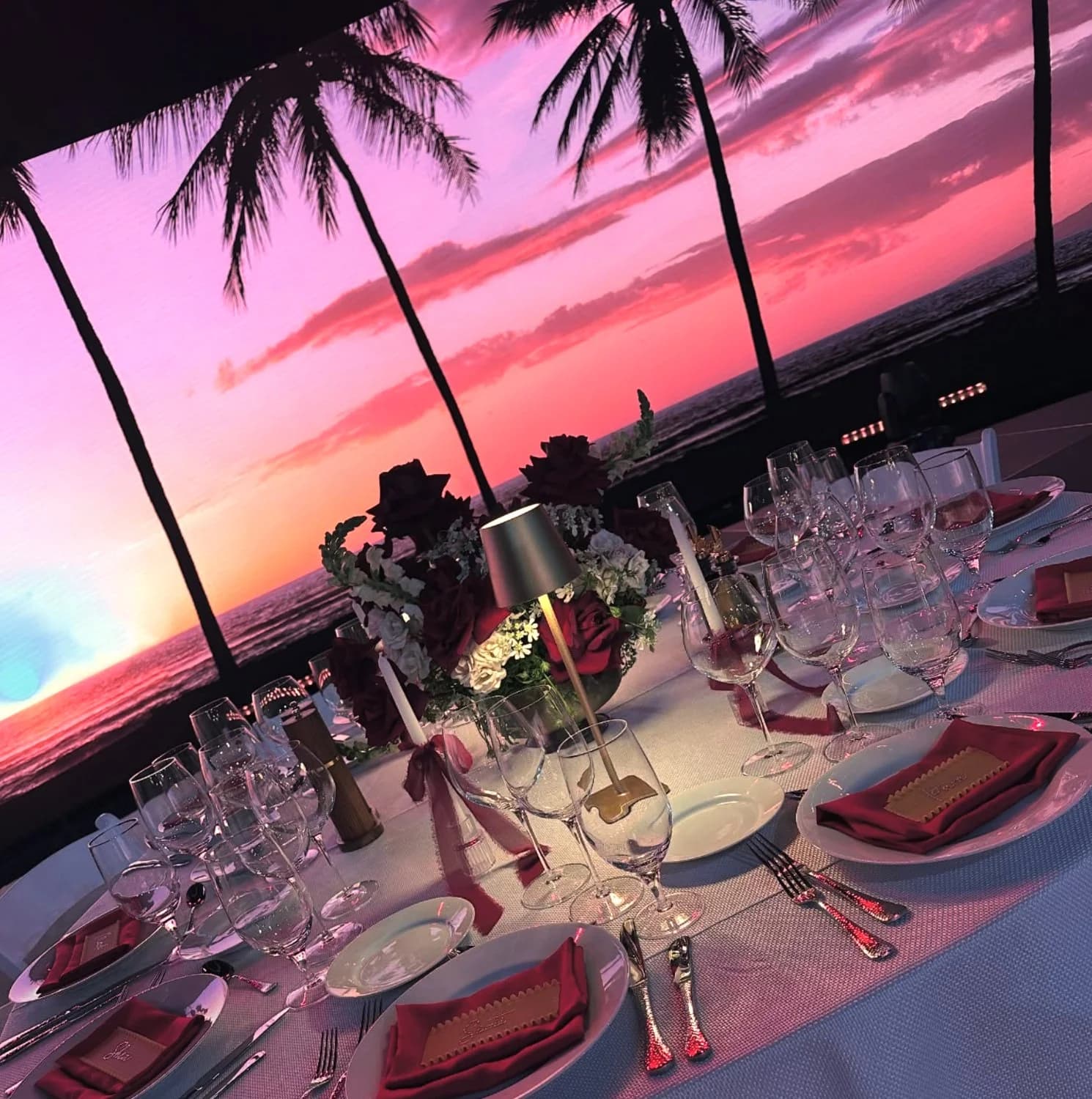 An intimate gala dinner table set with crystal and roses in front of the LED wall showing a tropical palm-lined sunset.