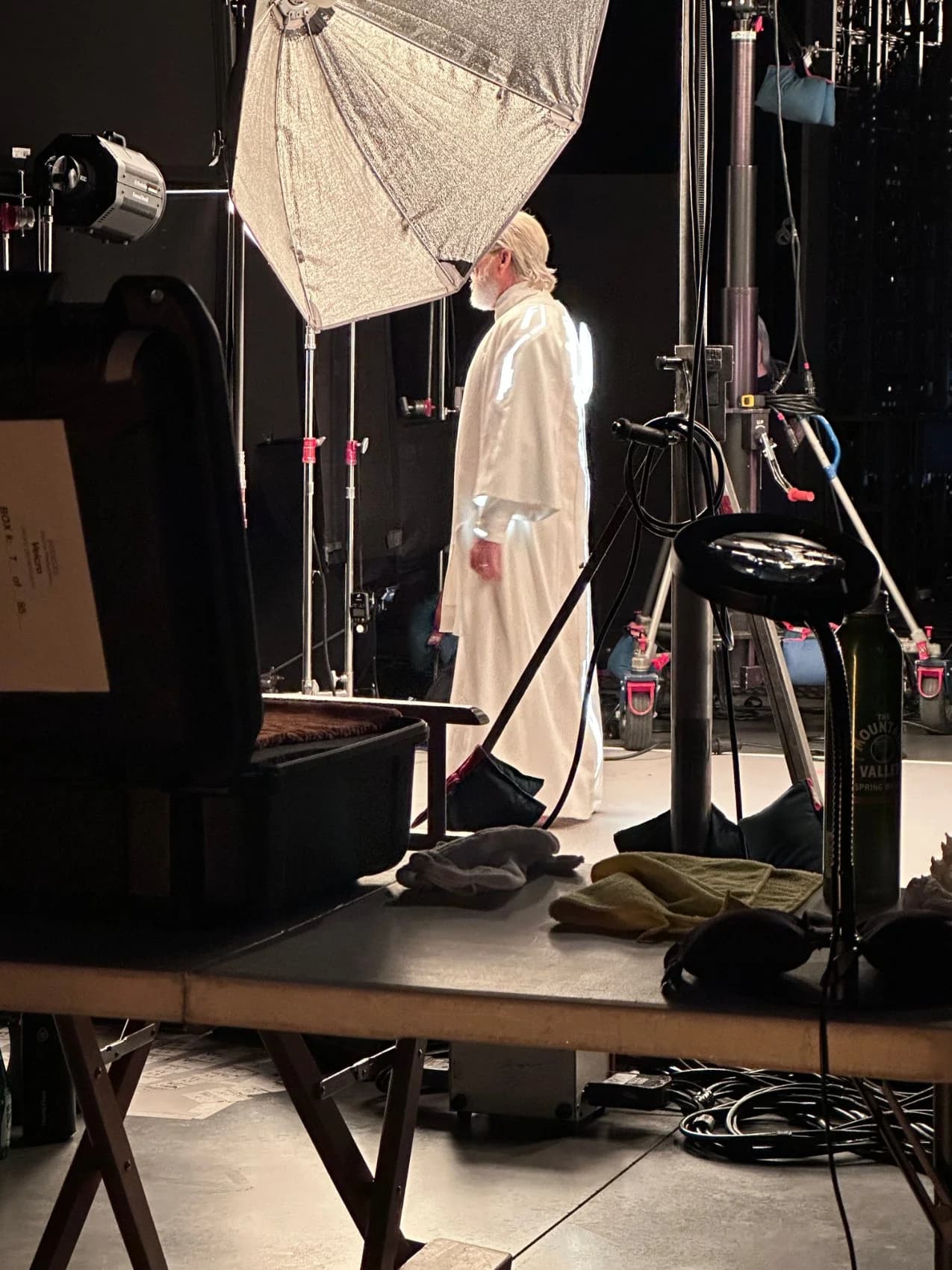 Back view of the illuminated white robe with a glowing emblem, the performer on the lit set surrounded by lighting gear.