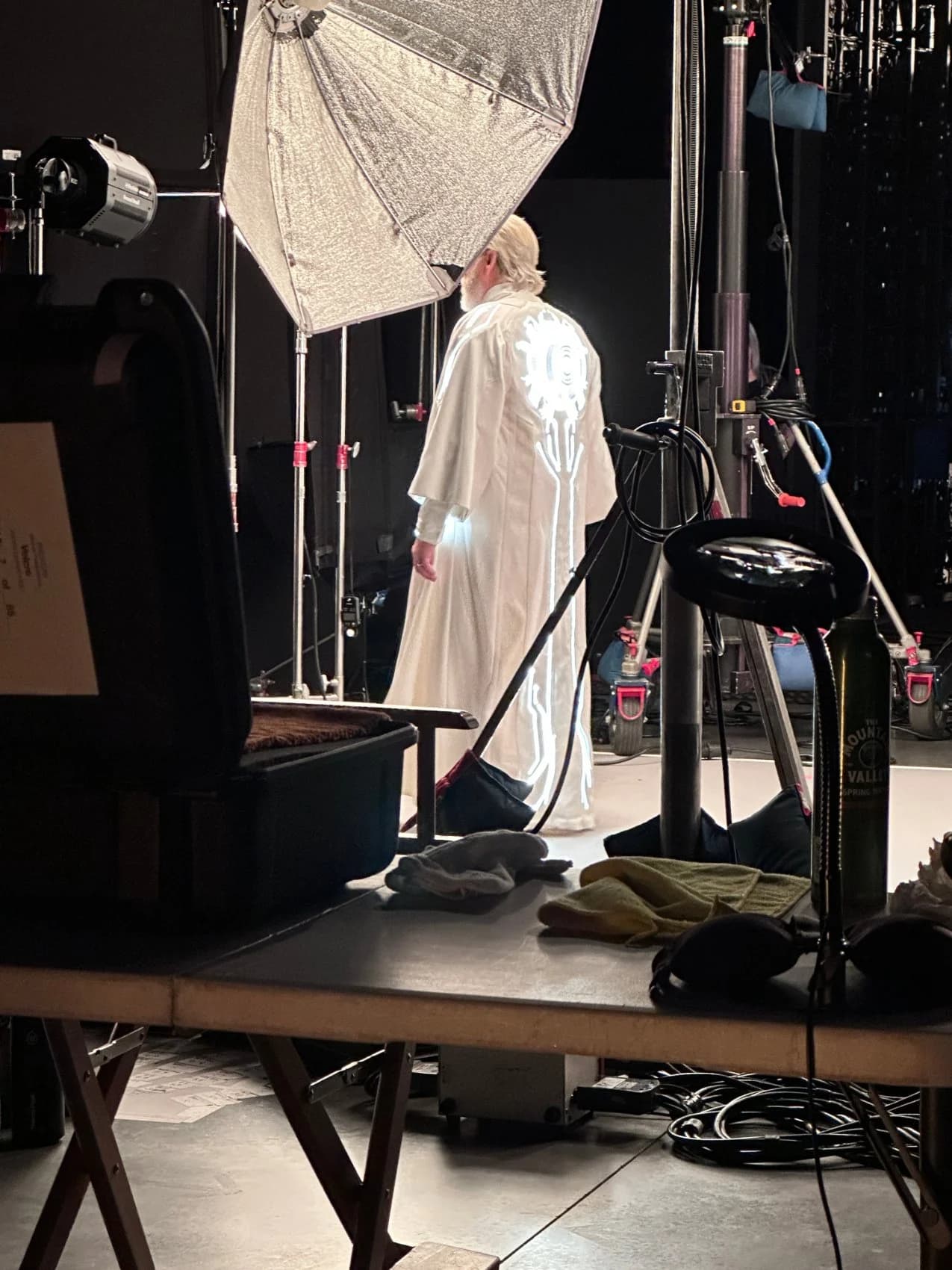 The glowing emblem on the back of the white robe as the performer faces the cyc wall, a crew hand adjusting a stand.