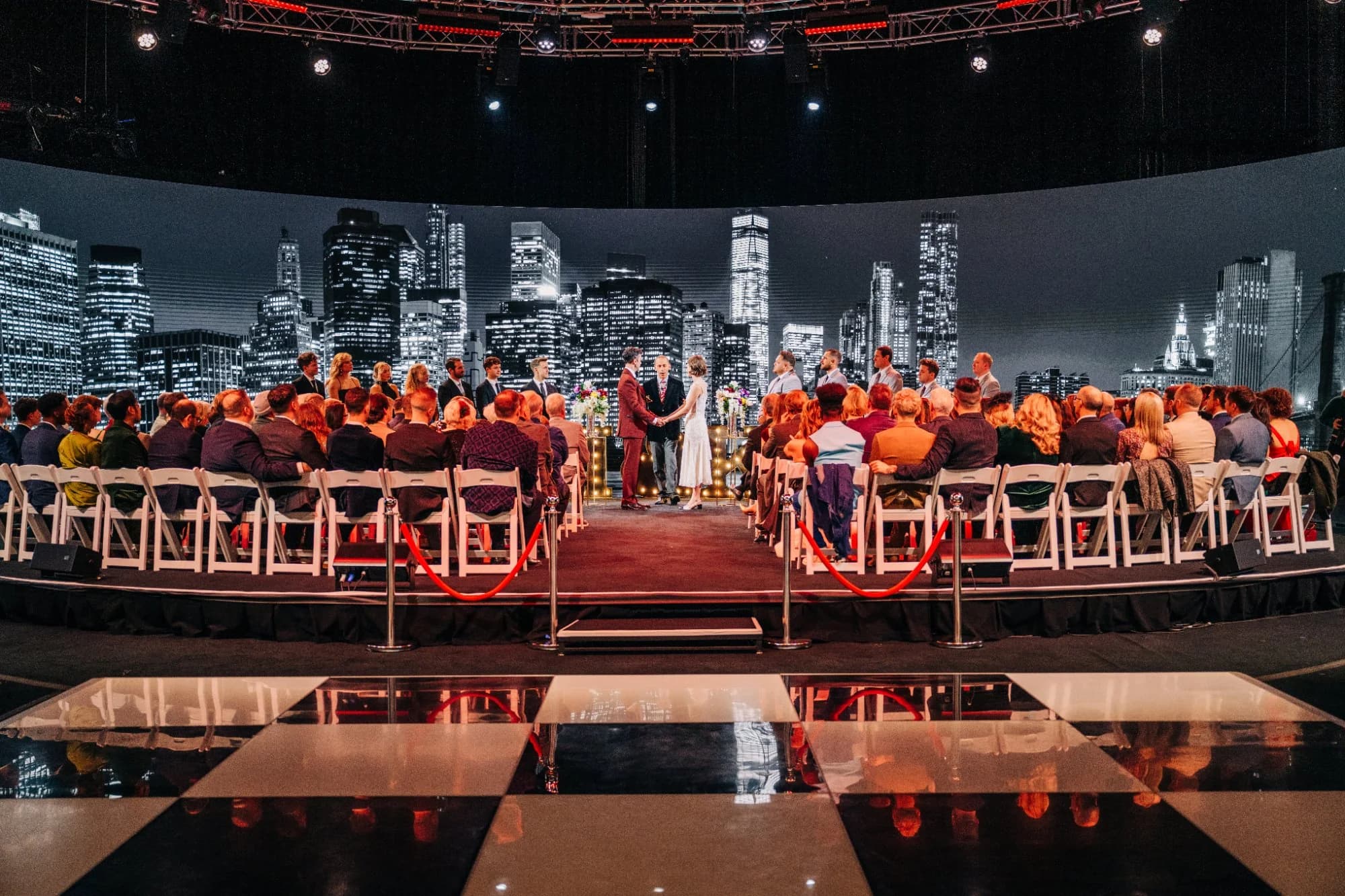 A wedding ceremony on the 360° stage at Worre Studios, guests seated along a red-carpet aisle as a glowing night-city skyline wraps the LED wall behind the couple.
