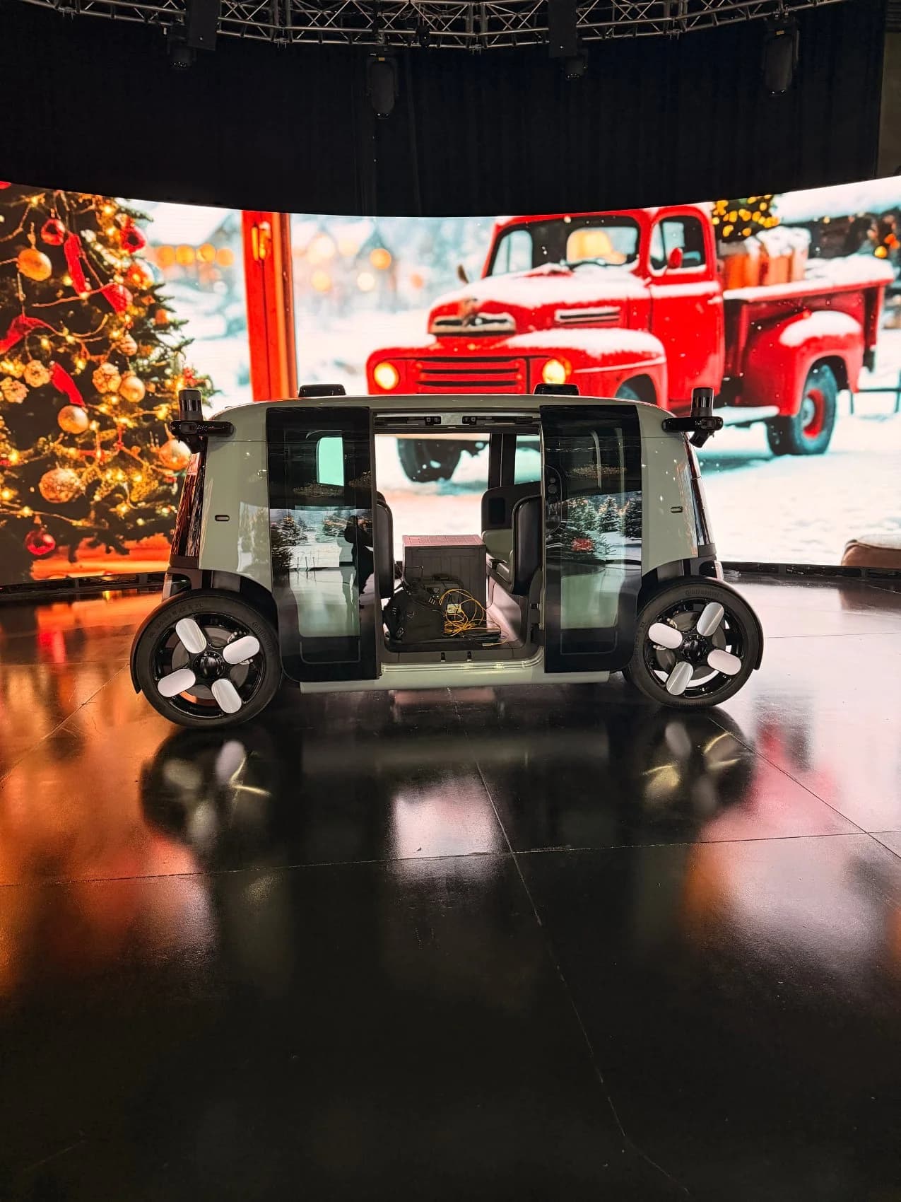 A Zoox robotaxi parked on the studio stage in front of a holiday scene — a snowy red pickup and lit Christmas tree — on the LED volume.