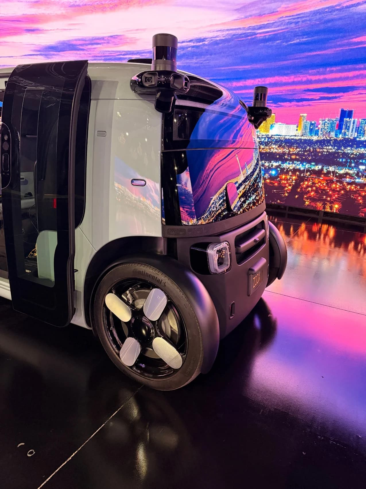 Close-up of a Zoox robotaxi on stage, its glossy body reflecting a Las Vegas skyline at sunset rendered across the LED wall.